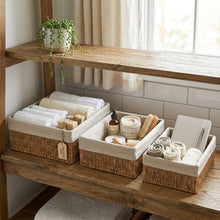 Drie open jute opbergmanden op een houten plank, gevuld met textiel en verzorgingsartikelen